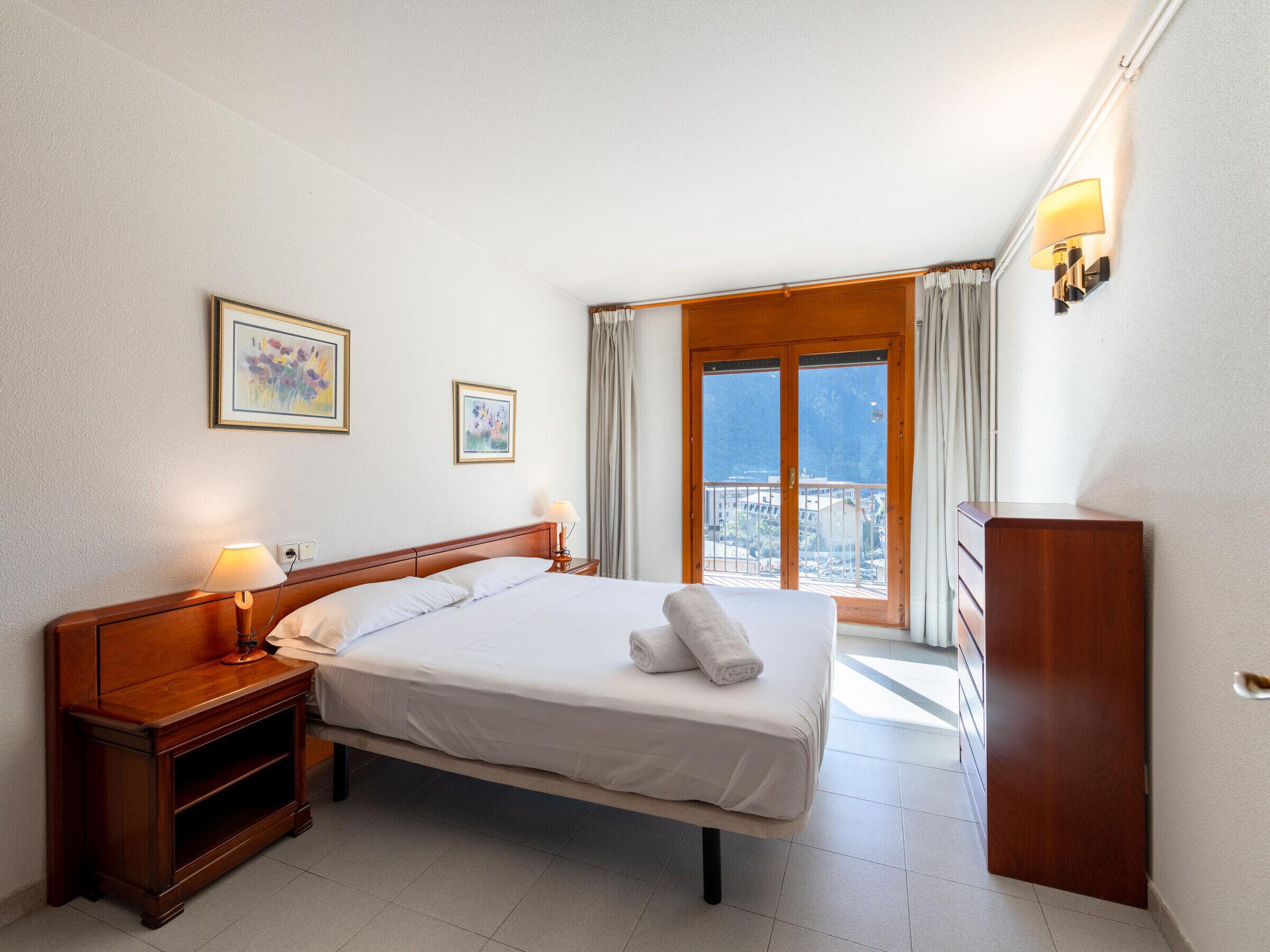 Een hotelkamer met een bed, nachtkastjes, een dressoir, een raam met gordijnen en twee ingelijste foto's aan de muur.