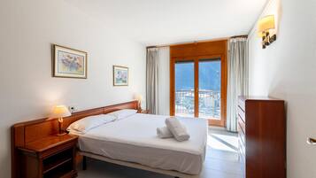 Een hotelkamer met een bed, nachtkastjes, een dressoir, een raam met gordijnen en twee ingelijste foto's aan de muur.