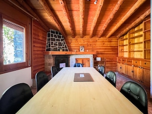 A wooden cabin interior with a long dining table, black leather chairs, a fireplace, and a stone-tiled chimney.