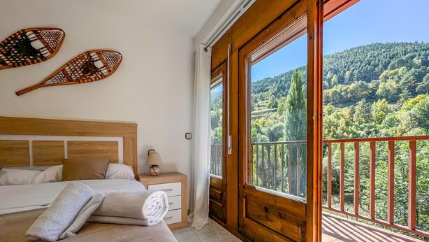 A bedroom with a large bed, a nightstand, a lamp, and a view of a forested hillside through a balcony. - Rider Apartment - 8 Guests, 4 Bedrooms (Pal)