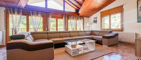 Ein geräumiges Wohnzimmer mit einem großen, abgewinkelten Ecksofa, einem Glastisch und einer Holzbalkendecke.