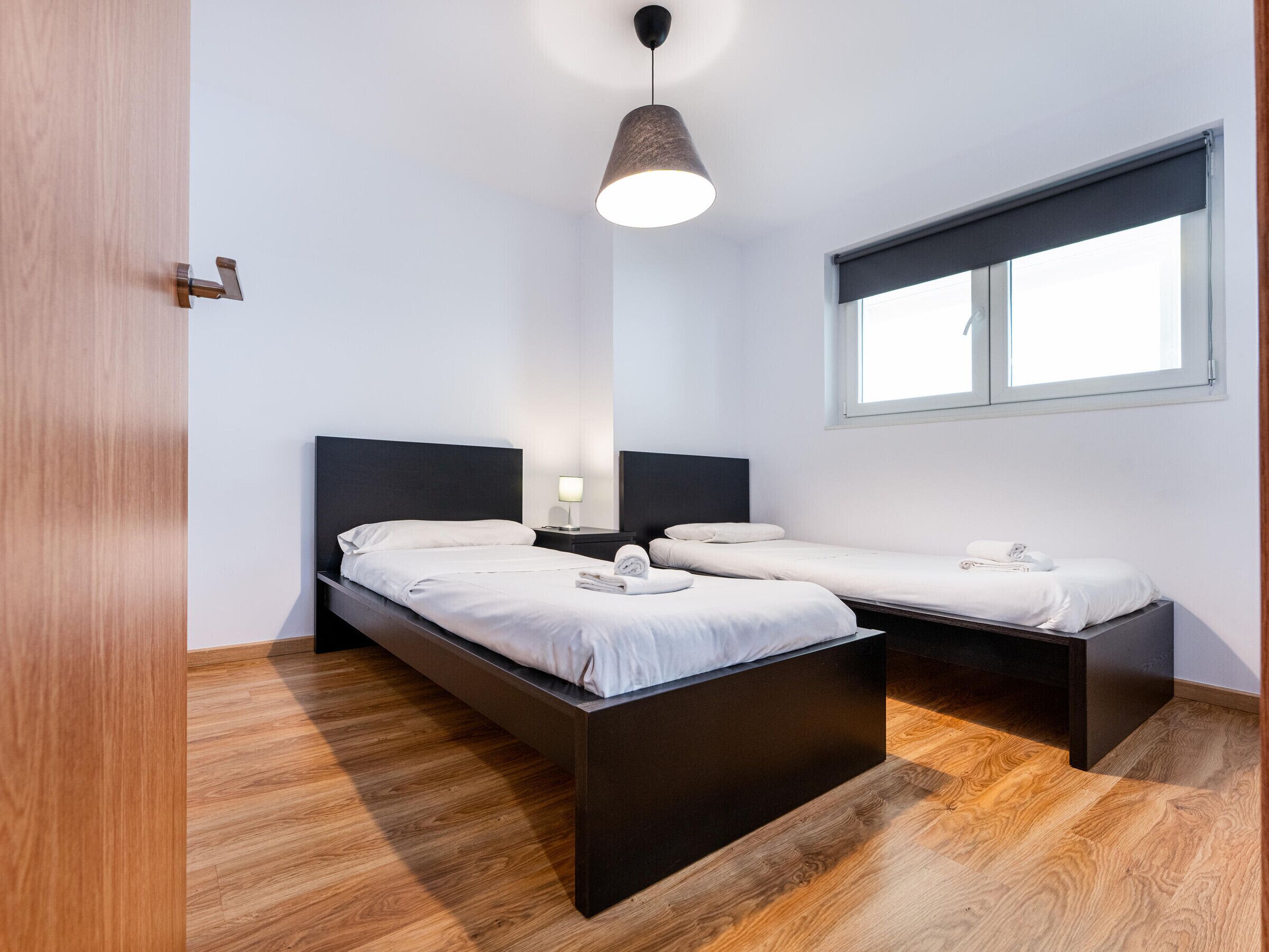 Een moderne slaapkamer met twee bedden, een houten vloer, een raam met zonwering en een hanglamp.