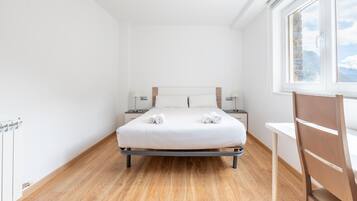 Ein modernes Schlafzimmer mit Holzboden, einem Bett mit weißen Leinen, zwei Nachttischen mit Lampen und einem Fenster mit Blick ins Freie.