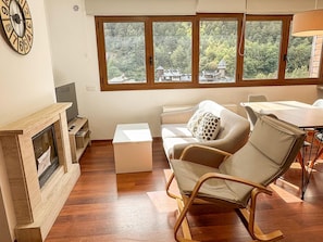 A living room with a fireplace, a sofa, a dining table, and a view of a forest through large windows.