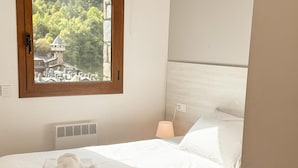 A neatly made bed with white linens and rolled towels, a bedside lamp, and a window with a view of a building and trees. - Modern Apartment, 5 ppl, 2 Bedrooms, Includes Parking (Arinsal)