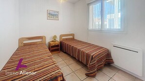 Un dormitorio con dos camas individuales, una mesita de noche, una lámpara, una luz en el techo, una ventana y un cuadro enmarcado en la pared.