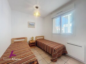 A bedroom with two single beds, a nightstand, a lamp, a ceiling light, a window, and a framed picture on the wall. - Practical apartment near the ocean - 4 guests (Vieux-Boucau-les-Bains)
