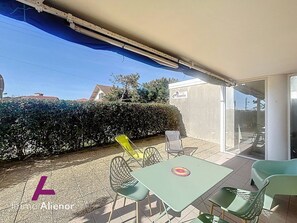 Un espace extérieur aménagé en terrasse, avec une table et des chaises, une chaise jaune, une haie et un bâtiment sur lequel on peut lire l’inscription « Atlantis ».
