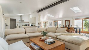 Uma sala de estar moderna com um sofá seccional, uma mesa de centro com livros e uma cozinha ao fundo.