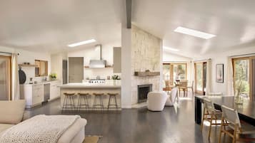 Uma cozinha moderna com ilha central, banquetas, lareira e área de jantar com luz natural.