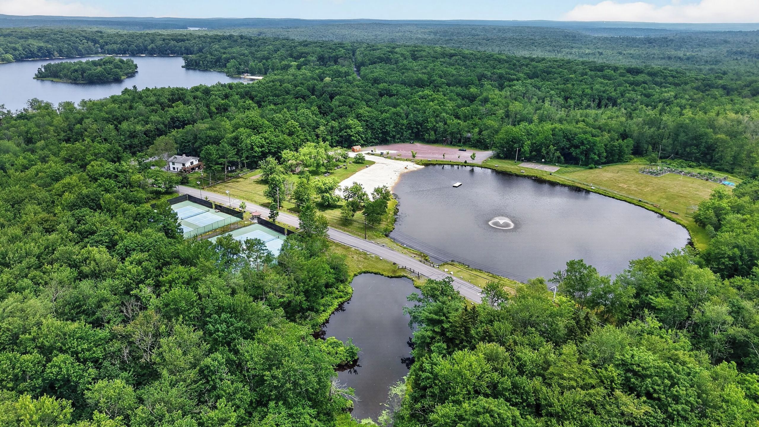 Vue aérienne d’une propriété au bord d’un lac, comprenant des courts de tennis, une piscine et des zones boisées environnantes.