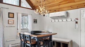 Et rustikt interiør med et træspisebord, stole, en lysekrone, et vindue med gardiner, vægmonterede kroge og en fastmonteret aircondition.