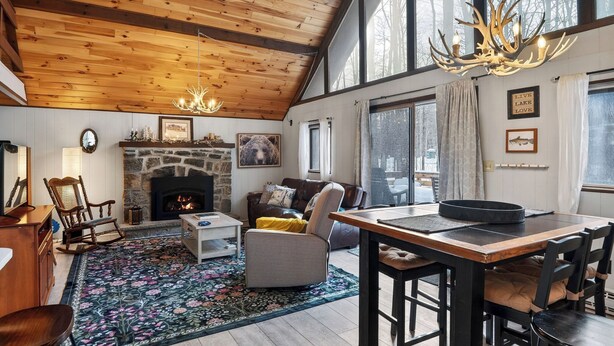 A cozy living room with a fireplace, a dining area with a table and chairs, and large windows with a view. - Evergreen Lodge by Avantstay Poconos Lake Cabin + Fireplace + Large Deck + Near Jack Frost & Kalahari (Pocono Lake)