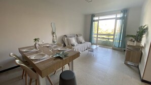 Un salón amplio con una mesa de comedor lista para cuatro comensales, un sofá, un balcón con vistas y una pequeña mesa con una planta.