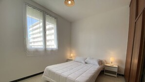 Un dormitorio con cama, dos mesitas de noche con lámparas, una ventana con persianas y una luz en el techo.