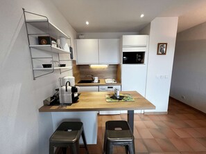 A compact kitchen with white cabinets, a coffee machine, a sink, and a small refrigerator. There are two stools and a shelf with kitchen items. - Charming apartment with parking - 4 guests (Port Grimaud)