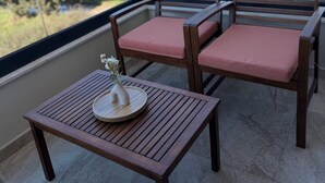 Un balcon avec deux chaises en bois et une petite table, un vase rempli de fleurs, et une vue sur les arbres et les bâtiments au loin.