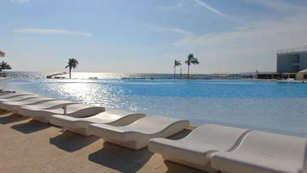 A poolside area with lounge chairs and palm trees, a clear sky, and a distant view of the sea. - Océano Aria, Entire Lodging: Condominium in Bucerias, Mexico (Nuevo Vallarta)