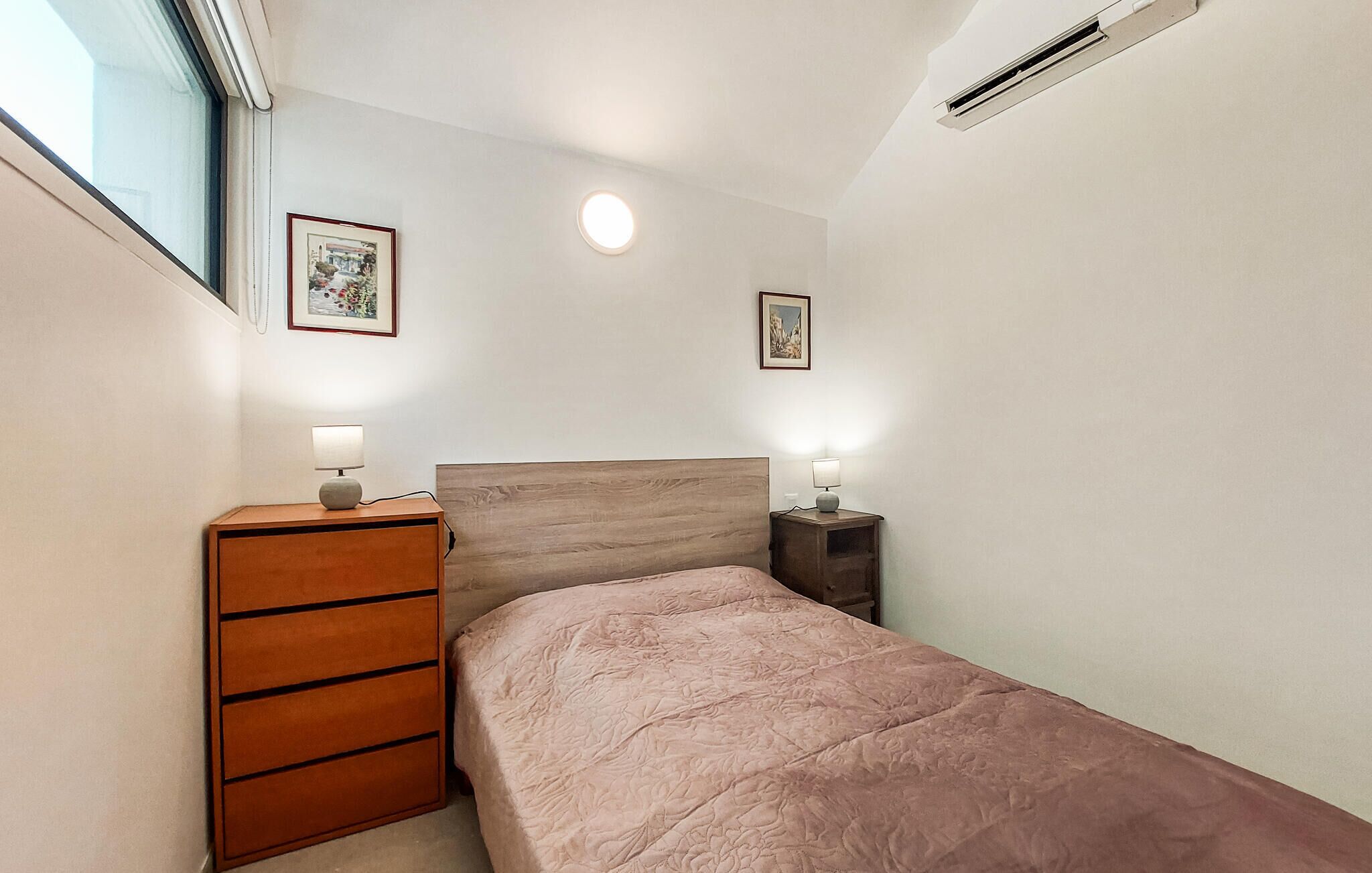 A bedroom with a bed, a dresser, two bedside tables with lamps, a window, and an air conditioning unit.