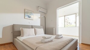 A modern bedroom with a large bed, a radiator, a ceiling fan, and a window with a view of buildings. - Terrace Dreams w/Pasalimani View - 2 minutes of walk distance (Pireas)