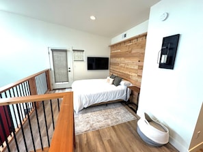 A modern bedroom with a wooden headboard, a bed with white bedding, a mounted TV, and a staircase with a wooden railing.