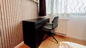 Une pièce avec un mur recouvert de lambris en bois, un bureau noir avec une chaise, un radiateur et une fenêtre ornée de rideaux à motifs.