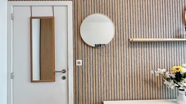 Une pièce avec un mur recouvert de lambris en bois, une porte blanche, un miroir rond, un meuble blanc à portes à persiennes et une petite étagère sur laquelle est posé un vase de fleurs.