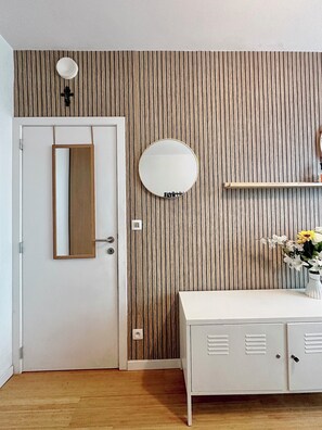 A room with a wooden slat wall, a white door, a round mirror, a white cabinet with louvered doors, and a small shelf with a vase of flowers. - Room for 3 people with private bathroom close to the mid station (Forest)