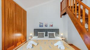 Un dormitorio con un amplio armario de madera, un cabecero tapizado en gris, dos mesitas de noche con lámparas y una escalera de madera.