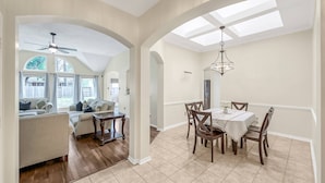 A spacious living area with a dining table and chairs, a ceiling fan, and large windows allowing natural light.