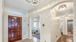 A spacious hallway with a wooden door, a chandelier, and a view into a room with a desk and chair.