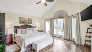 A bedroom with a large bed, a ceiling fan, a wooden floor, a window with curtains, and a TV mounted on the wall.