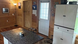 A wooden cabin interior with a granite countertop, a white door, a small window, and a cabinet with pots. - Charming 1-bedroom house on 2 acres in peaceful Orrington (Orrington)