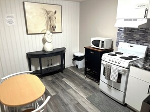 A compact kitchen with a microwave, stove, and a trash bin. There's a wooden chair and a table, and a framed picture of a horse on the wall.