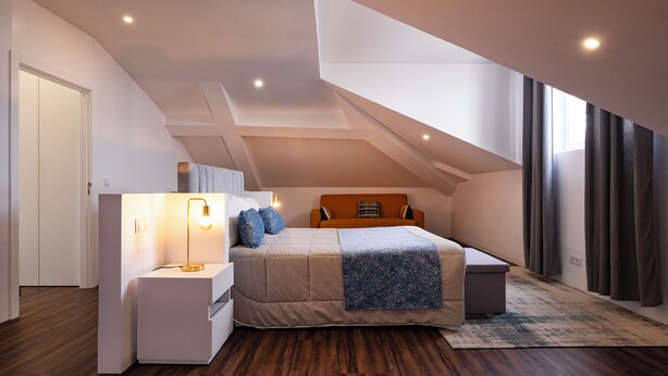A modern bedroom with a sloped ceiling, a bed with a beige bedspread, a bedside table with a lamp, and a large window with curtains. - Snuggly Corner by Trip2Portugal (Figueira da Foz)
