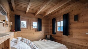 A wooden cabin room with a bed, two windows, a small table, and a lamp. - Chalet Uska - Enjoy the thrill of the Alps all year round - OVO Network (Combloux)