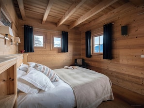 Una habitación de cabaña de madera con una cama, dos ventanas, una mesita y una lámpara.
