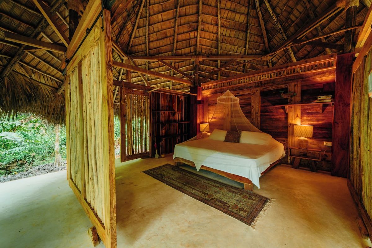 A wooden room with a bed, a nightstand, and a thatched roof.