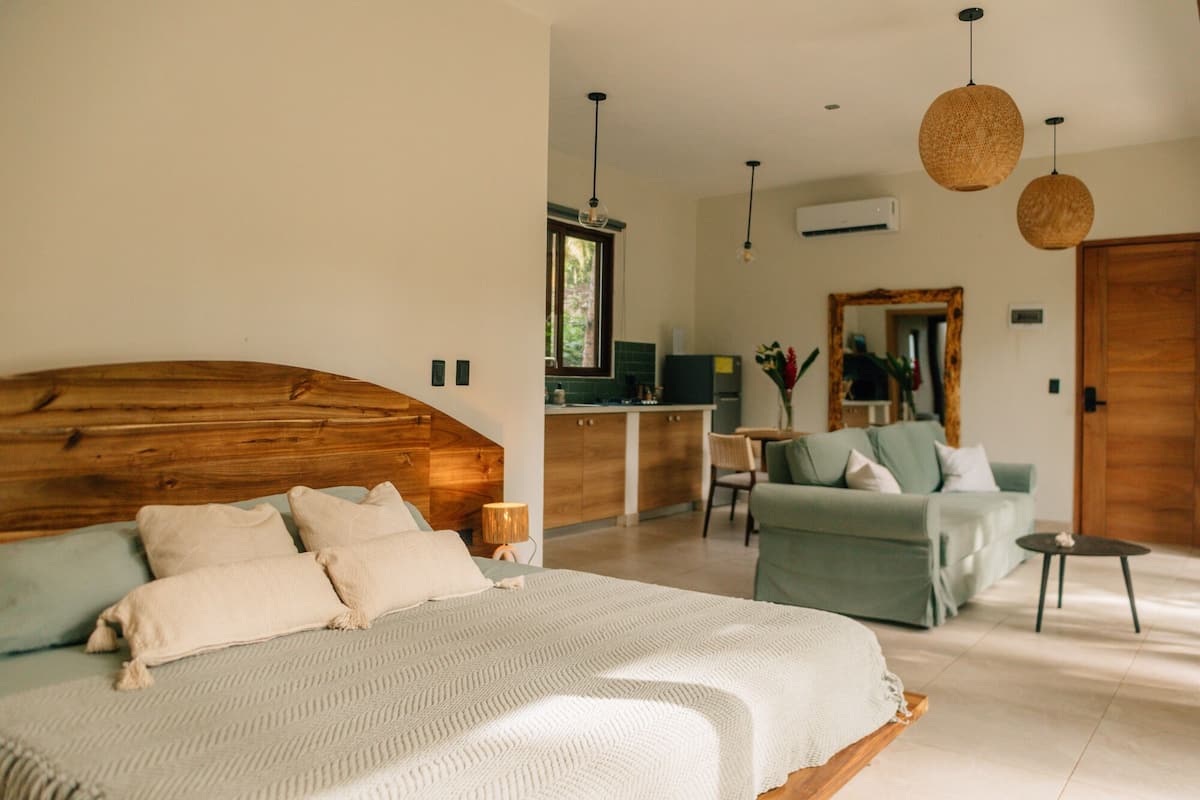 A bedroom with a wooden headboard, a large bed with a white blanket, a desk with a computer, a green sofa, and a dining table.
