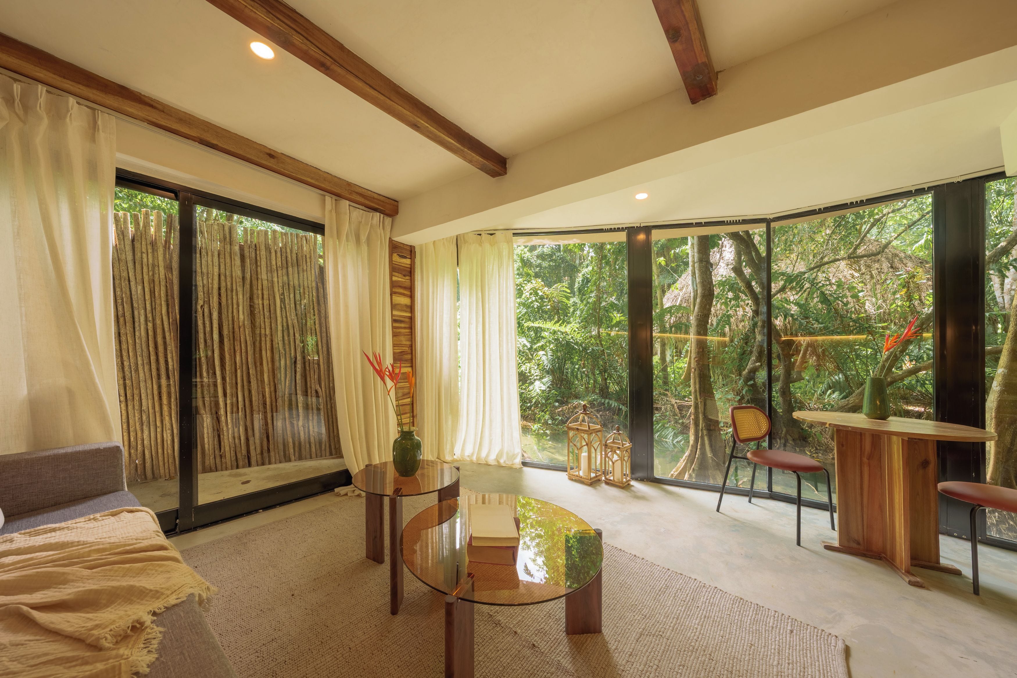 A modern living room with a glass coffee table, a sofa, and a view of a lush garden through large windows.