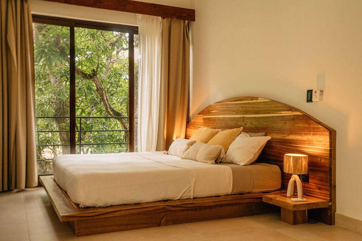 A bedroom with a large window, a wooden headboard, a bedside lamp, and a bed with white linens.