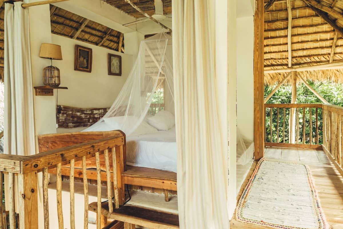 A wooden bed with a canopy, a small table with a lamp, and a view of greenery through a balcony.