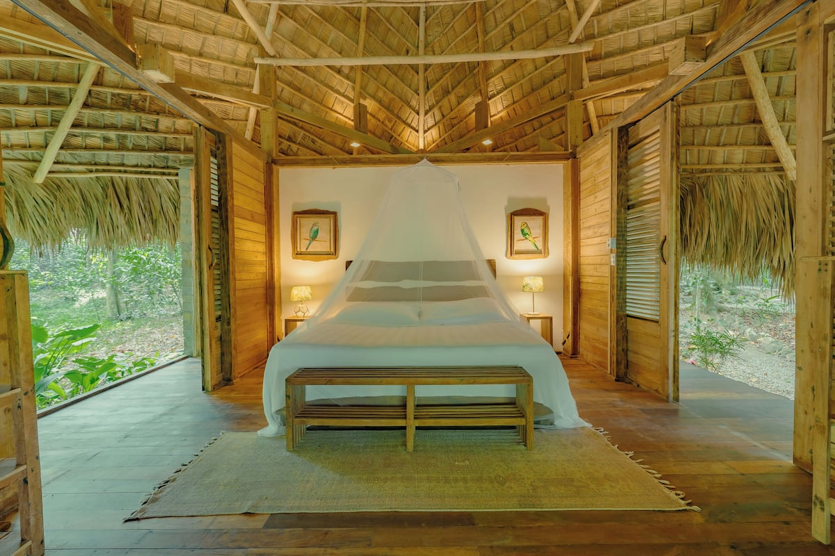 A room with a bed, wooden bench, framed pictures, and a thatched roof.