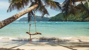 Una playa con agua turquesa cristalina, una hamaca colgando de una palmera y un paisaje montañoso al fondo.