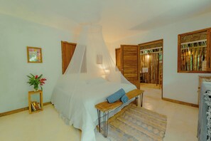 A bedroom with a canopy bed, a bedside table, a bench, a bedside lamp, a framed picture, and a door leading to another room.