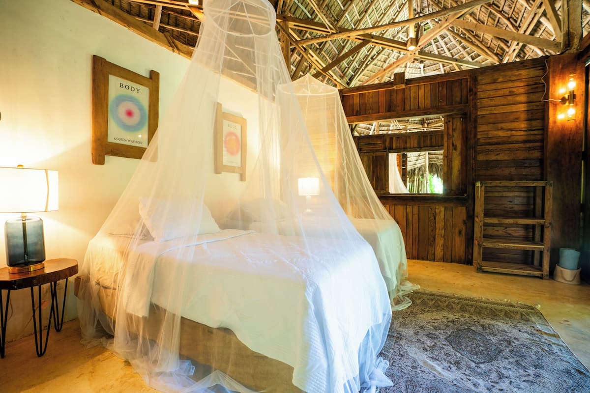 A room with a bed under a canopy, a wooden wall, a framed picture, a lamp, and a window.