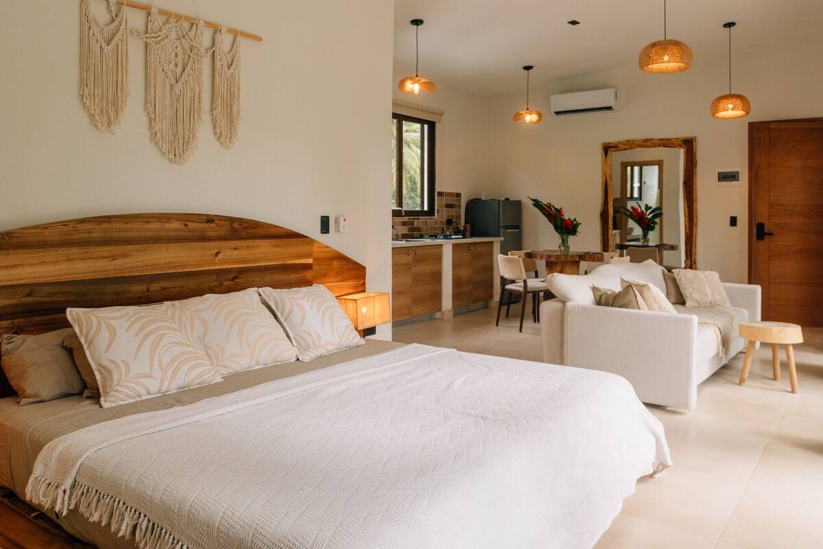 A bedroom with a wooden headboard, a large bed, a sitting area with a sofa and chairs, a small table, and a kitchen area with a refrigerator and stove.