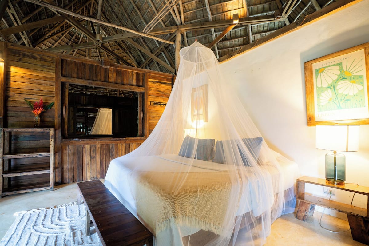 A room with a bed under a canopy, wooden walls, a bench, a lamp, and a framed picture on the wall.