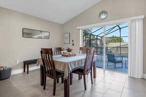 A dining area with a table set for four, surrounded by wooden chairs. A large window offers a view of an outdoor swimming pool and patio furniture. - Spacious 3 bedroom,2 bathroom family home with private pool (Davenport)