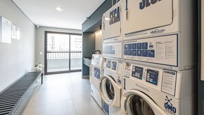 A laundry area with multiple washing machines, a bench, and a view of the outside through a glass door. - Studio 407 Perdizes 900m Allianz Parque c Academia (São Paulo)